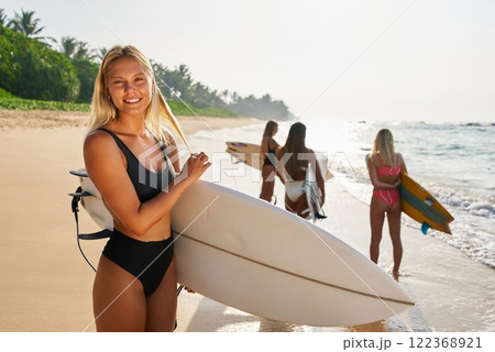 Women in swimsuits carry surfboards at tropical beach. Blonde holds board, ready to surf. Friends in ocean waves, summer lifestyle. Palm trees. Surf culture, ocean vibes, active holiday. 122368921
