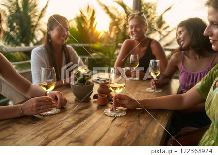 Women enjoy evening wine in tropical restaurant. Friends laugh, share stories, celebrate bachelorette party traveling. Relaxed meeting of diverse women. Sunset scenery adds warm glow. Women enjoy evening wine in tropical restaurant. Friends laugh, share stories, celebrate bachelorette party traveling. Relaxed meeting of diverse women. Sunset scenery adds warm glow. 122368924