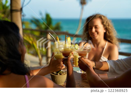 Women toast drinks at tropical beachside restaurant. Friends celebrate bachelorette party trip. Cheers with cocktails in summer. Diverse group enjoying vacation. 122368925
