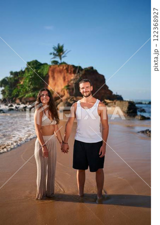 Couple holding hands on sandy beach, smiles facing camera. Man in tank, woman in dress enjoy romantic walk by sea. Sunset lights cliff backdrop. Honeymooners by ocean, travel love concept. 122368927