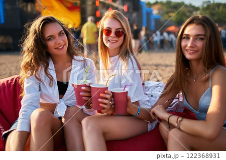 Group of girls relax on red bean bag chairs, enjoying drinks at sunny beach music festival. Three young females in summer attire, smiling, chilling waterfront event. Audience at coastal gig, sunset. 122368931