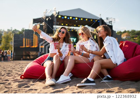 Group of female friends enjoy summer music festival on sandy beach, sitting on red bean bag chairs, sipping cool drinks, stage in background. Happy women in casual attire relax, laugh. Group of female friends enjoy summer music festival on sandy beach, sitting on red bean bag chairs, sipping cool drinks, stage in background. Happy women in casual attire relax, laugh. 122368932