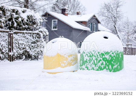 Garbage sorting bins covered with snow on winter day, fence and grey house in a background 122369152