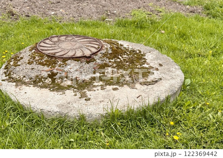 A rusty iron hatch with a pattern covered dirt above an observation well is located next to a green grass in the daytime. Close-up A rusty iron hatch with a pattern covered dirt above an observation well is located next to a green grass in the daytime. Close-up 122369442