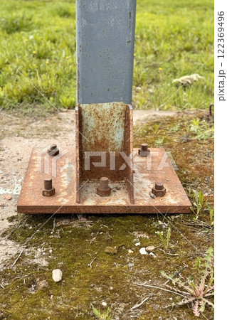 Metal pillar fastened to basement with anchor bolts and nuts. The rusty surface of the post and the fastenings are in places. The support of the iron black pillar on a green lawn in the park Metal pillar fastened to basement with anchor bolts and nuts. The rusty surface of the post and the fastenings are in places. The support of the iron black pillar on a green lawn in the park 122369496