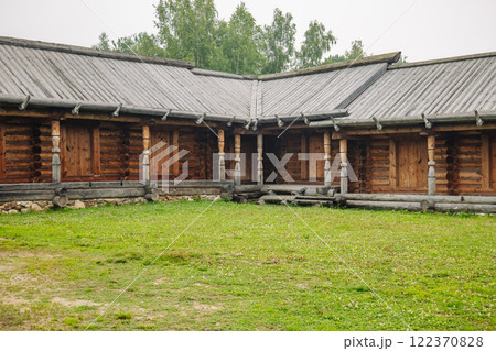 Log cabin with porch and carved pillars stands on green grass field in a cloudy day, representing russian rural architecture 122370828