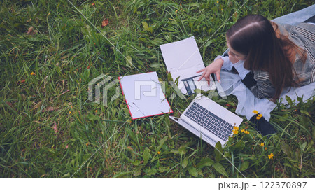 Schoolgirl lying on lush grass, using laptop and calculator while completing homework outdoors, enjoying the peaceful autumn day Schoolgirl lying on lush grass, using laptop and calculator while completing homework outdoors, enjoying the peaceful autumn day 122370897