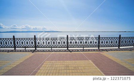 Paved riverbank walkway featuring decorative metal railing, stretching alongside tranquil water under bright azure skies, presenting serene urban landscape 122371075