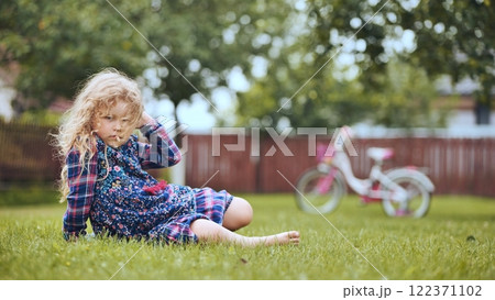 Blonde girl sitting on grass in backyard, touching her hair, with bicycle in background, enjoying a peaceful family day Blonde girl sitting on grass in backyard, touching her hair, with bicycle in background, enjoying a peaceful family day 122371102