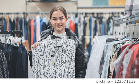 Smiling shopper gripping shirt hanger, browsing clothing store filled with vibrant fashion selections Smiling shopper gripping shirt hanger, browsing clothing store filled with vibrant fashion selections 122371118