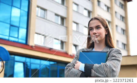 Young student holding folders in her arms, standing in front of university building, looking away with thoughtful expression Young student holding folders in her arms, standing in front of university building, looking away with thoughtful expression 122371261