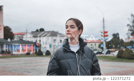 Stylish young woman wearing black puffy jacket and white turtleneck enjoys a cloudy day in a town square 122371272