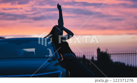 Carefree woman silhouetted against sunset, leaning on car after driving through scenic landscape 122371276