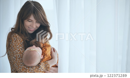 Young mother cradling her sleeping newborn baby girl near a window with white curtains, enjoying a peaceful moment at home 122371289