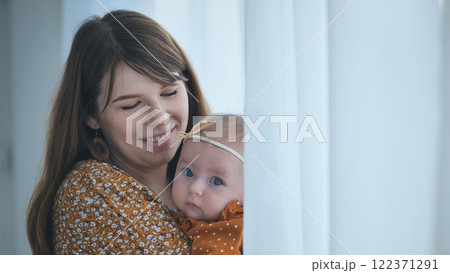 Radiant mother embracing newborn near sunlit window, soft curtains casting gentle illumination across tender bonding moment 122371291