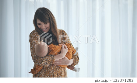 Smiling mother holding her sleeping baby girl by the window at home, enjoying motherhood and childcare Smiling mother holding her sleeping baby girl by the window at home, enjoying motherhood and childcare 122371292