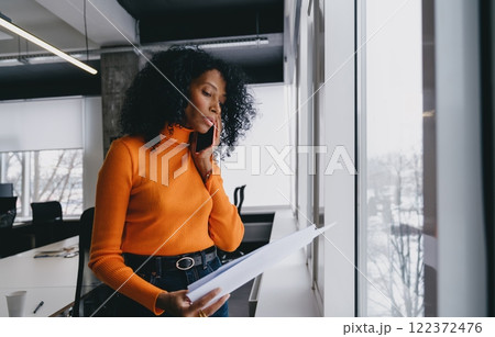 A focused businesswoman in an orange sweater engages in a critical phone call while analyzing a document, discussing a trending eco-strategy, snowy cityscape visible through the office window. A focused businesswoman in an orange sweater engages in a critical phone call while analyzing a document, discussing a trending eco-strategy, snowy cityscape visible through the office window. 122372476