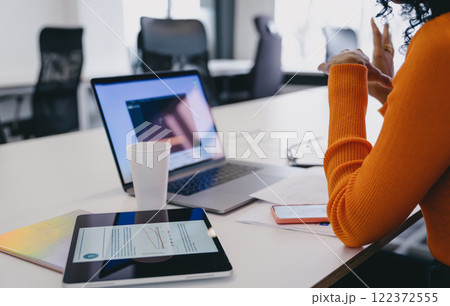 African American entrepreneur analyzing market trends on tablet at co-working, with laptop and smartphone, in orange sweater, embodying essence of private enterprise and agile business management. 122372555
