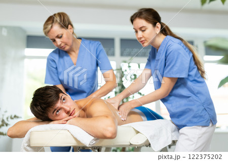 Two women doing back massage to man lying on massage table in spa salon Two women doing back massage to man lying on massage table in spa salon 122375202