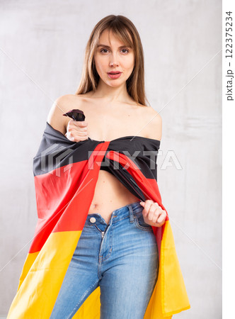 Flirting young woman in jeans with state flag of Germany against gray wall indoors Flirting young woman in jeans with state flag of Germany against gray wall indoors 122375234