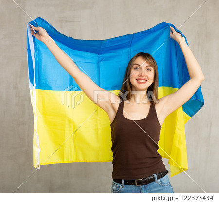 Portrait of confident smiling young female student standing against gray wall with national flag of Ukraine 122375434