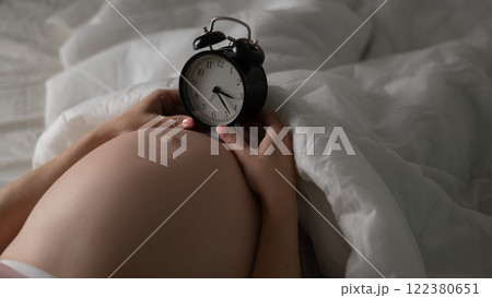 A pregnant woman lies in bed and holds an alarm clock. Close up of the belly. A pregnant woman lies in bed and holds an alarm clock. Close up of the belly. 122380651