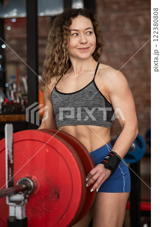Caucasian forty year old woman putting a weight plate on a barbell in the gym. Vertical photo.  122380808