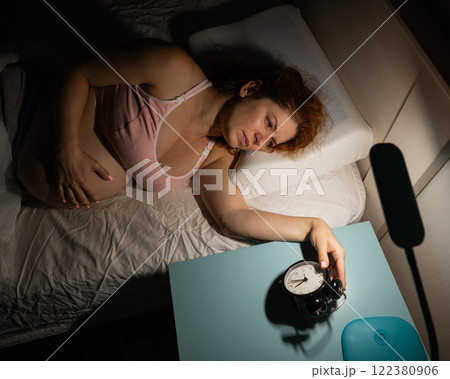 A pregnant woman lies in bed and holds an alarm clock.  122380906