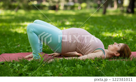 Pregnant caucasian woman doing yoga in the park. Prenatal shoulder bridge exercise.  122381011