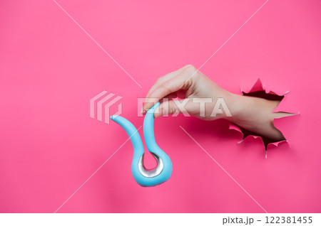Female hand sticking out of pink paper background with blue curved vibrator sex toy. Copy space. Female hand sticking out of pink paper background with blue curved vibrator sex toy. Copy space. 122381455