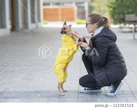 Young Caucasian woman in a jacket walks with a small dog in overalls.  122381528