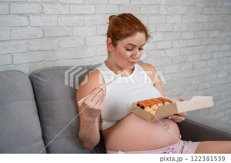 A pregnant woman sits on the sofa and eats rolls from a box. Food delivery.  122381539