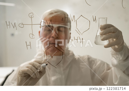 An elderly Caucasian male scientist in a protective suit holds a test tube and writes a formula on the glass. An elderly Caucasian male scientist in a protective suit holds a test tube and writes a formula on the glass. 122381714