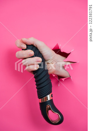 Female hand sticking out of pink paper background with black textured vibrator. Vertical photo. Female hand sticking out of pink paper background with black textured vibrator. Vertical photo. 122381791