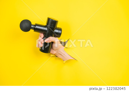 Female hand sticking out with massage gun from yellow paper background. Copy space.  122382045