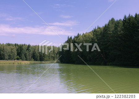 Picturesque view of a forest lake with clear water reflecting green trees and blue sky. The shore is covered with dense coniferous and deciduous trees. Film photography effect and grunge effect 122382203