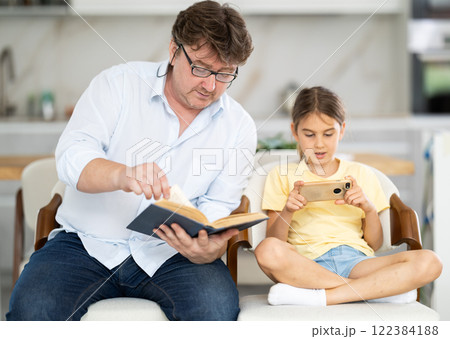 Father reading book, daughter using phone Father reading book, daughter using phone 122384188