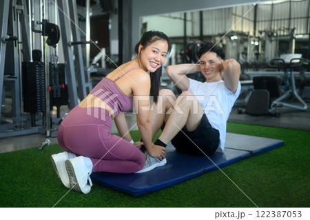 Smiling young woman assisting her workout partner by holding his feet during sit-ups on a gym mat 122387053