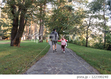 A young girl smiles brightly, holding a stuffed animal, while walking with her father in a green 122390240