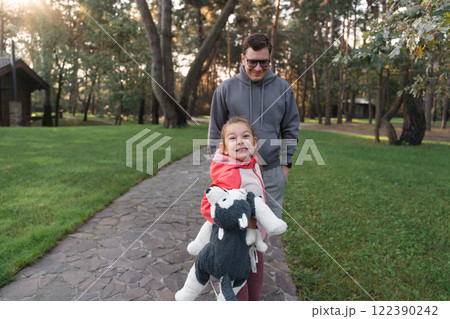 A young girl smiles brightly, holding a stuffed animal, while walking with her father in a green 122390242