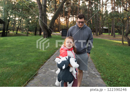 A young girl smiles brightly, holding a stuffed animal, while walking with her father in a green 122390246