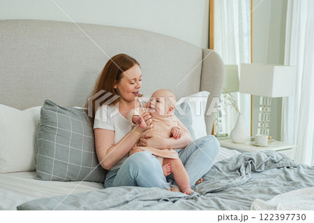 Happy family at home. Mother playing with little toddler child on bed. Mom baby relax playing having fun together. Mother looks at baby with love care. Mom of breast feeding baby rest in living room Happy family at home. Mother playing with little toddler child on bed. Mom baby relax playing having fun together. Mother looks at baby with love care. Mom of breast feeding baby rest in living room 122397730