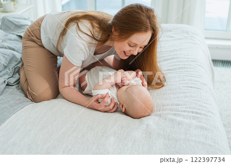 Happy family at home. Mother playing with little toddler child on bed. Mom baby relax playing having fun together. Mother looks at baby with love care. Mom of breast feeding baby rest in living room Happy family at home. Mother playing with little toddler child on bed. Mom baby relax playing having fun together. Mother looks at baby with love care. Mom of breast feeding baby rest in living room 122397734