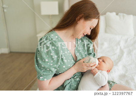 Happy family at home. Mother holding little baby toddler child feeding milk from bottle. Newborn Infant baby girl sucking eating drinking milk. Mom of breast feeding baby. Motherhood happy child 122397767