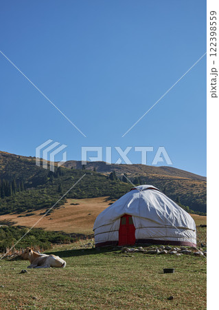 Serene Yurt Positioned Amidst Nature, Complemented by a Vast Grassland Background. cow is lying nearby beautiful, picturesque yurt is set amidst a lush, green landscape, all under vast, clear blue sky 122398559