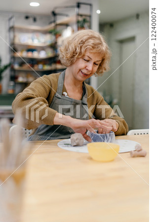 Elderly Caucasian woman sculpts clay bowl at pottery workshop, learning new skill. Senior lady enjoys working with hands, developing creativity and fine motor skills Elderly Caucasian woman sculpts clay bowl at pottery workshop, learning new skill. Senior lady enjoys working with hands, developing creativity and fine motor skills 122400074