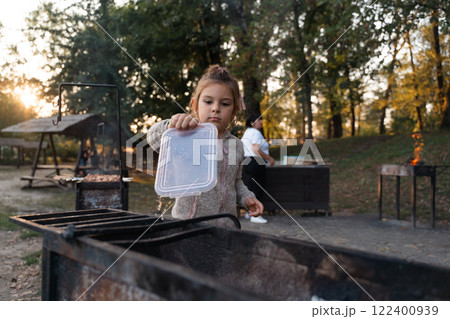 A young girl assists with a barbecue by holding a container over the grill, surrounded by trees and 122400939