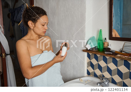 Woman standing in bathroom examining cosmetic bottle wrapped in towel Woman standing in bathroom examining cosmetic bottle wrapped in towel 122401134