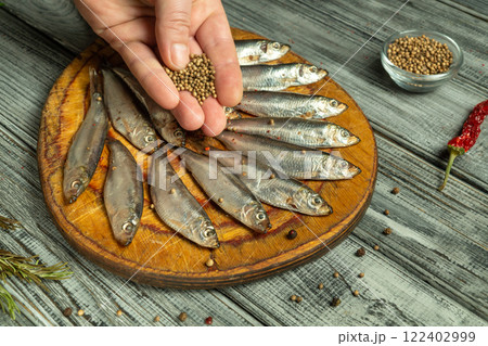 A hand delicately sprinkles aromatic spices or coriander on a round wooden board adorned with freshly washed fish. The rustic kitchen setting evokes culinary charm, attracting the taste buds A hand delicately sprinkles aromatic spices or coriander on a round wooden board adorned with freshly washed fish. The rustic kitchen setting evokes culinary charm, attracting the taste buds 122402999