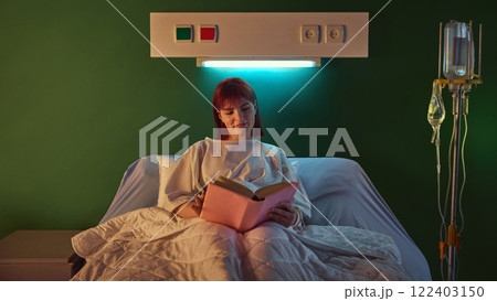 Positive and relaxed young woman in hospital gown lying in bed and reading book in the evening after her procedures in well-equipped hospital ward in clinic 122403150
